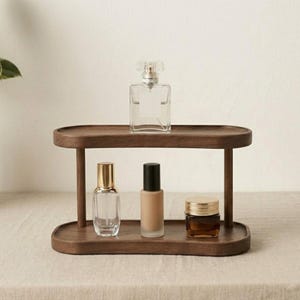 May include: A two-tiered, dark wood display stand holding various beauty products. The top shelf features a clear perfume bottle with a decorative stopper. The bottom shelf holds a perfume bottle, a foundation bottle, and a jar with a gold lid.