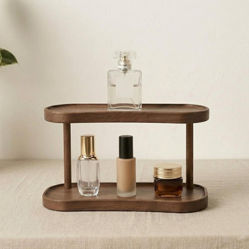 May include: A two-tiered, dark wood display stand holding various beauty products. The top shelf features a clear perfume bottle with a decorative stopper. The bottom shelf holds a perfume bottle, a foundation bottle, and a jar with a gold lid.