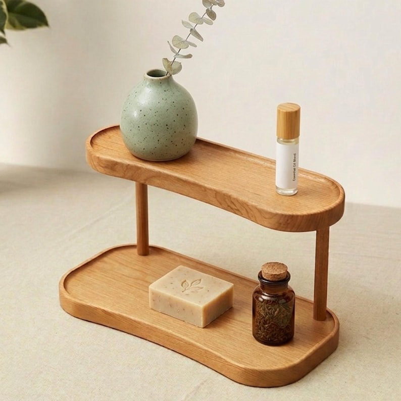 May include: A two-tiered wooden tray with rounded edges, holding a small green vase with a sprig of greenery, a roller bottle, a bar of soap, and a small glass bottle with a cork. The tray is made of light-colored wood.