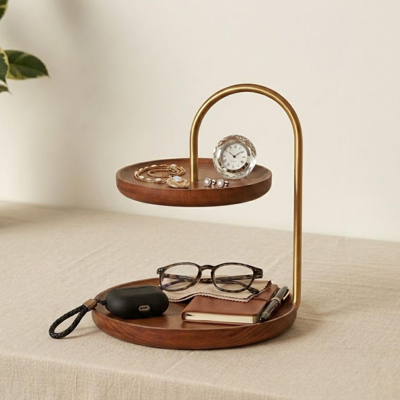 Puede incluir: Un organizador de madera de dos niveles con un marco de metal dorado. El nivel superior contiene un peque&ntilde;o reloj redondo, una pulsera dorada y pendientes. El nivel inferior contiene unas gafas de carey, un estuche negro para auriculares y una libreta.