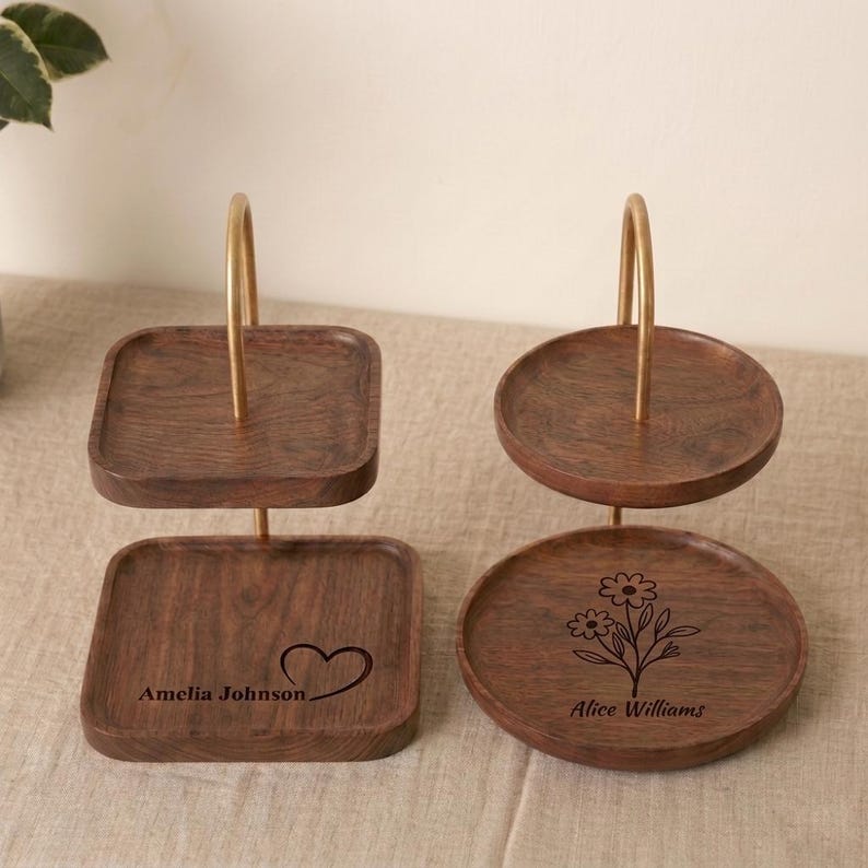 Puede incluir: Dos soportes de madera de dos niveles con asas de metal dorado. Uno tiene una base cuadrada grabada con el nombre Amelia Johnson y un coraz&oacute;n. El otro tiene una base redonda grabada con una ilustraci&oacute;n de una flor y el nombre Alice Williams.