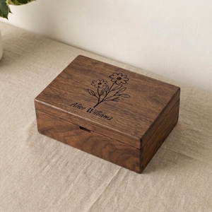 Puede incluir: Una caja rectangular de madera marrón oscuro con tapa con bisagras. La caja presenta un diseño floral grabado y el nombre "Alice Williams". La caja está sobre una superficie beige.