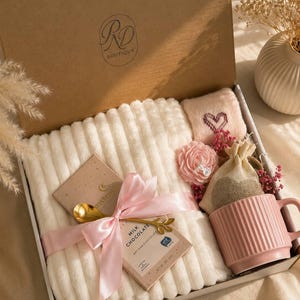 Peut inclure: Un coffret cadeau contenant une couverture blanche c&ocirc;tel&eacute;e, un mug rose &agrave; stries verticales, une tablette de chocolat au lait nou&eacute;e d'un ruban rose et d'une cuill&egrave;re dor&eacute;e, une petite pochette rose brod&eacute;e d'un c&oelig;ur, une fleur et un sac en toile de jute.