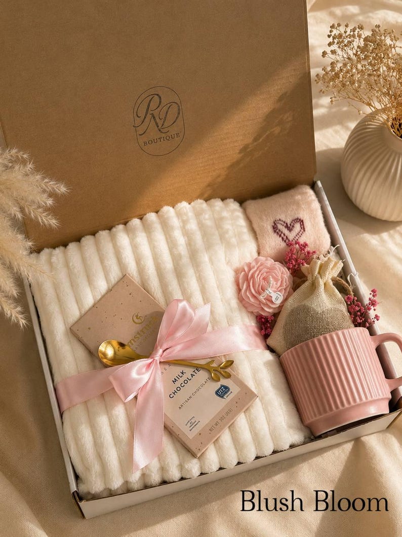 Peut inclure: Un coffret cadeau contenant une couverture blanche c&ocirc;tel&eacute;e, un mug rose &agrave; stries verticales, une tablette de chocolat au lait nou&eacute;e d'un ruban rose et d'une cuill&egrave;re dor&eacute;e, une petite pochette rose brod&eacute;e d'un c&oelig;ur, une fleur et un sac en toile de jute.