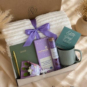 Peut inclure: Un coffret cadeau contenant un plaid blanc c&ocirc;tel&eacute; avec un ruban violet, un carnet vert avec un stylo dor&eacute;, une tablette de chocolat noir Moonstruck, un tube en verre de sels de bain violets, une tasse verte c&ocirc;tel&eacute;e et un petit bouquet avec une &eacute;tiquette Best Wishes.