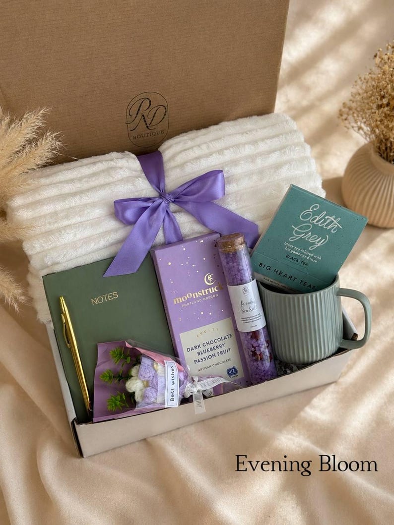 Peut inclure: Un coffret cadeau contenant un plaid blanc c&ocirc;tel&eacute; avec un ruban violet, un carnet vert avec un stylo dor&eacute;, une tablette de chocolat noir Moonstruck, un tube en verre de sels de bain violets, une tasse verte c&ocirc;tel&eacute;e et un petit bouquet avec une &eacute;tiquette Best Wishes.