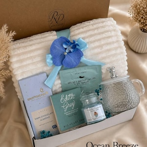 Peut inclure: Un coffret cadeau contenant une couverture blanche c&ocirc;tel&eacute;e nou&eacute;e d'un ruban bleu et d'une orchid&eacute;e, une tablette de chocolat noir aux amandes Moonstruck, un masque pour les yeux &agrave; l'acide hyaluronique Peauamie, du th&eacute; noir Edith Grey, une bougie Vanilla Cupcake et une tasse en verre avec couvercle.