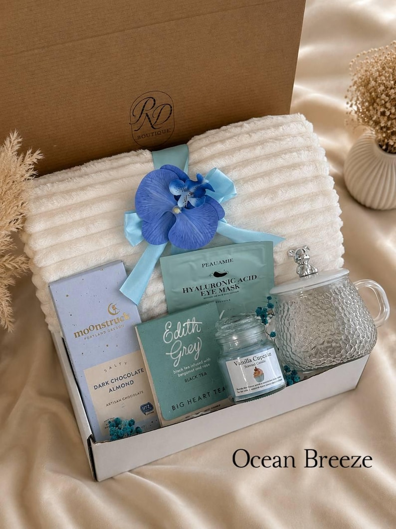 Peut inclure: Un coffret cadeau contenant une couverture blanche c&ocirc;tel&eacute;e nou&eacute;e d'un ruban bleu et d'une orchid&eacute;e, une tablette de chocolat noir aux amandes Moonstruck, un masque pour les yeux &agrave; l'acide hyaluronique Peauamie, du th&eacute; noir Edith Grey, une bougie Vanilla Cupcake et une tasse en verre avec couvercle.