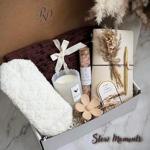 May include: A gift box filled with self-care items. Includes a brown blanket, fluffy white socks, a candle, a notebook with a gold pen, a flower-shaped hair clip, and a small bottle of pink sea salt. The box has the text "Slow Moments".