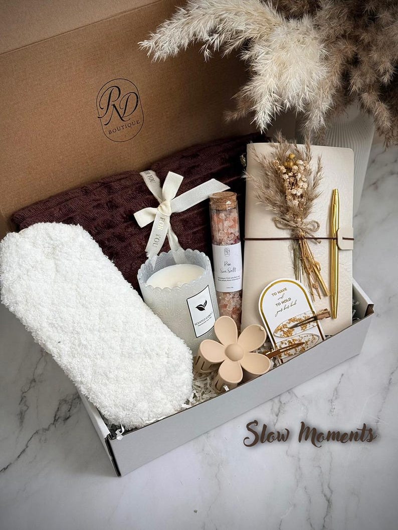May include: A gift box filled with self-care items. Includes a brown blanket, fluffy white socks, a candle, a notebook with a gold pen, a flower-shaped hair clip, and a small bottle of pink sea salt. The box has the text "Slow Moments".