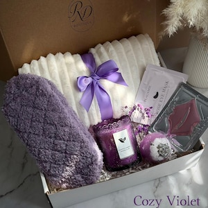 May include: A gift box labeled "RD Boutique" filled with self-care items. Includes a white plush blanket tied with a purple ribbon, a purple eye mask, a scented candle, bath bomb, and rose eye mask. The box is labeled "Cozy Violet".
