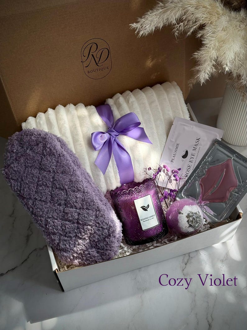 May include: A gift box labeled "RD Boutique" filled with self-care items. Includes a white plush blanket tied with a purple ribbon, a purple eye mask, a scented candle, bath bomb, and rose eye mask. The box is labeled "Cozy Violet".