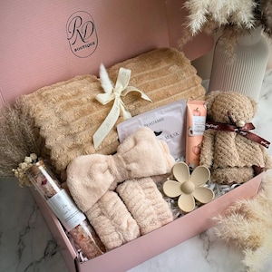 May include: A pink gift box filled with self-care items. Includes a plush towel, headband, wristbands, face mask, lotion, a teddy bear, and a flower-shaped hair clip. The box also contains a glass tube of bath salts. The text "Soft Hug" is visible.