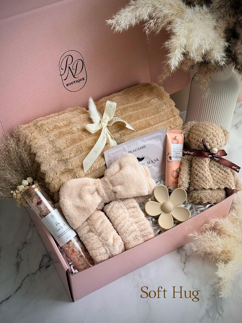 May include: A pink gift box filled with self-care items. Includes a plush towel, headband, wristbands, face mask, lotion, a teddy bear, and a flower-shaped hair clip. The box also contains a glass tube of bath salts. The text "Soft Hug" is visible.