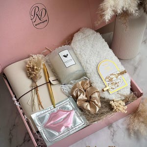 May include: A pink gift box labeled "RD Boutique" contains a notebook, pen, candle, face mask, hair scrunchies, and a fluffy white towel. The box also includes a gold hair clip and dried flowers. The text "Calm Elegance" is printed on the box.