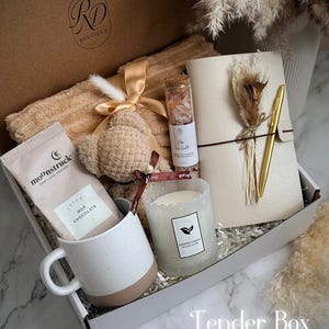 May include: A gift box with a variety of items, including a beige blanket, a teddy bear, a mug, a candle, a notebook, a pen, and a chocolate bar. The box is labeled "Tender Box."