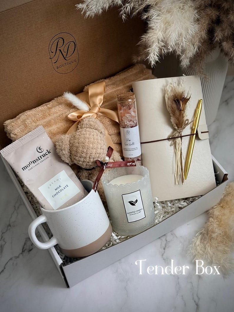 May include: A gift box with a variety of items, including a beige blanket, a teddy bear, a mug, a candle, a notebook, a pen, and a chocolate bar. The box is labeled "Tender Box."