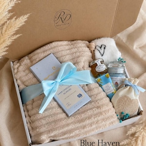 Peut inclure: Un coffret cadeau contenant un plaid beige textur&eacute; nou&eacute; avec un ruban bleu clair, une tablette de chocolat noir aux amandes, un petit pot de miel avec une cuill&egrave;re en bois, une bougie parfum&eacute;e, une cr&egrave;me pour les mains et un petit sachet.