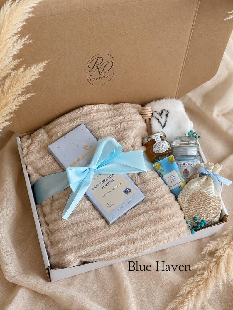 Peut inclure: Un coffret cadeau contenant un plaid beige textur&eacute; nou&eacute; avec un ruban bleu clair, une tablette de chocolat noir aux amandes, un petit pot de miel avec une cuill&egrave;re en bois, une bougie parfum&eacute;e, une cr&egrave;me pour les mains et un petit sachet.