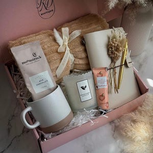May include: A pink gift box filled with a beige blanket, a white and brown speckled mug, a packet of cocoa, a candle, a notebook, a pen, and a tube of hand cream. The box is labeled "Soft Peach."