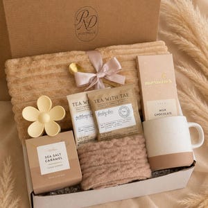 Peut inclure: Un coffret cadeau contenant une couverture côtelée beige avec un ruban rose, une pince à cheveux fleur jaune, deux sachets de thé Tea With Tae, une tablette de chocolat au lait Moonstruck, des biscuits au caramel et un mug en céramique.