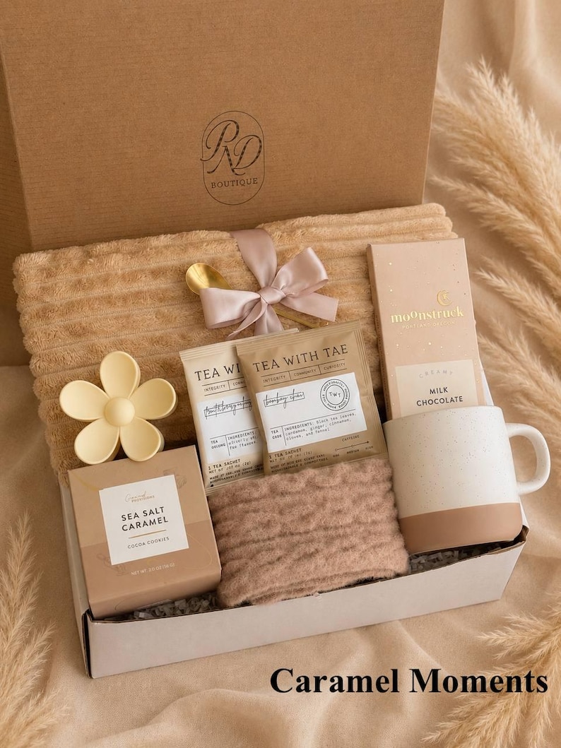 Peut inclure: Un coffret cadeau contenant une couverture c&ocirc;tel&eacute;e beige avec un ruban rose, une pince &agrave; cheveux fleur jaune, deux sachets de th&eacute; Tea With Tae, une tablette de chocolat au lait Moonstruck, des biscuits au caramel et un mug en c&eacute;ramique.