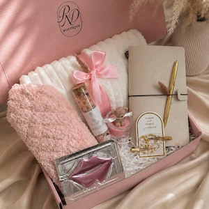 Peut inclure: Une bo&icirc;te cadeau rose contenant une couverture blanche textur&eacute;e nou&eacute;e d'un ruban rose, une paire de chaussettes duveteuses roses, un flacon en verre de sels de bain, une bougie avec un ours, un carnet beige, un stylo dor&eacute; et deux pinces &agrave; cheveux.