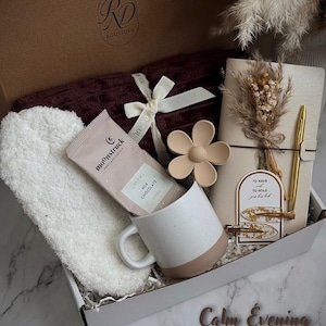 May include: A gift box with a burgundy blanket, white headband, a mug with a pink base, a chocolate bar, a flower-shaped object, a notebook, and a gold pen. The box is labeled "RD Boutique" and includes the text "Calm Evening".