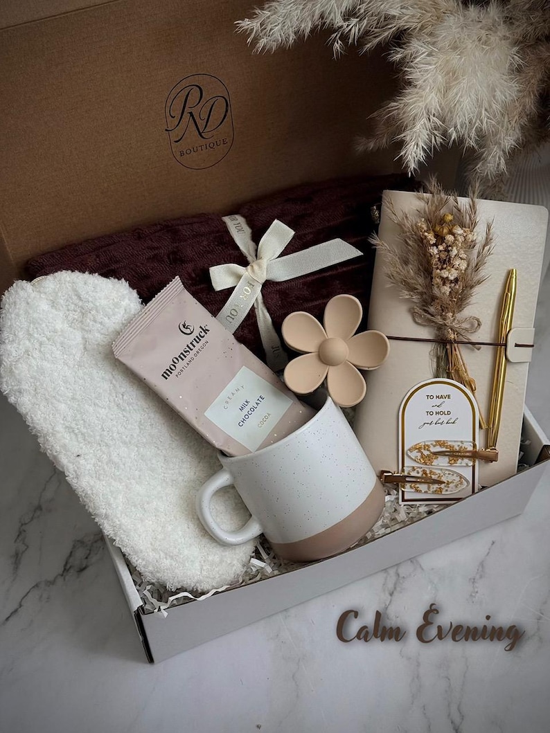 May include: A gift box with a burgundy blanket, white headband, a mug with a pink base, a chocolate bar, a flower-shaped object, a notebook, and a gold pen. The box is labeled "RD Boutique" and includes the text "Calm Evening".