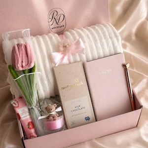 Peut inclure: Un coffret rose contenant une couverture en peluche blanche, une tulipe rose, une barre de chocolat, un carnet, un stylo, un petit ours en peluche et une crème pour les mains. Le coffret porte les mentions "RD Boutique" et "Soft Petals".