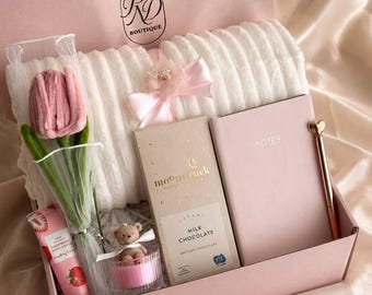 May include: A pink gift box filled with a white plush blanket, a pink tulip, a chocolate bar, a notebook, a pen, a small teddy bear, and a hand cream. The box has the text "RD Boutique" and "Soft Petals".