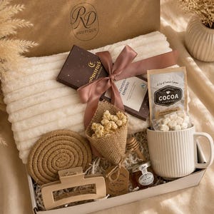 Peut inclure: Un coffret cadeau contenant un plaid blanc c&ocirc;tel&eacute;, une tablette de chocolat noir avec un ruban mauve, un sachet de cacao saveur vanille fran&ccedil;aise, une tasse blanche c&ocirc;tel&eacute;e, un petit pot de miel, une cuill&egrave;re &agrave; miel en bois, une pince &agrave; cheveux beige et des fleurs s&eacute;ch&eacute;es.