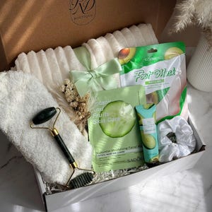 May include: A gift box labeled "RD Boutique" filled with self-care items. Includes a white fluffy towel, a green jade roller, face masks, a scrunchie, and a tube of avocado oil. The box is tied with a light green ribbon. Text on the box reads "Green Ritual".
