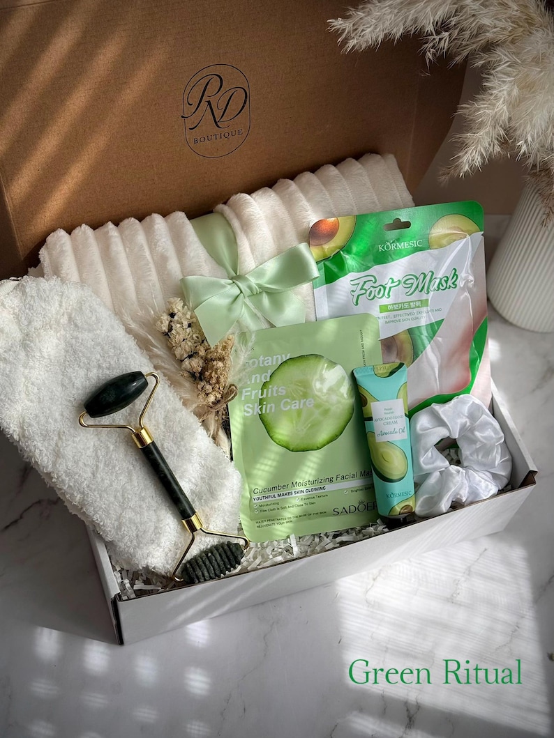 May include: A gift box labeled "RD Boutique" filled with self-care items. Includes a white fluffy towel, a green jade roller, face masks, a scrunchie, and a tube of avocado oil. The box is tied with a light green ribbon. Text on the box reads "Green Ritual".