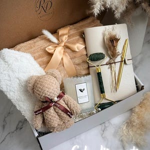May include: A gift box with a teddy bear, a candle, a face roller, a notebook, and a pen. The box also contains a towel and a ribbon. The text "Calm Moments" is written on the bottom of the image.