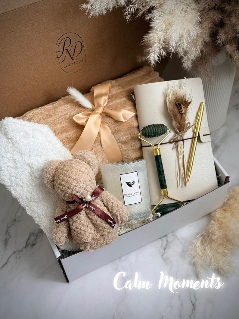 May include: A gift box with a teddy bear, a candle, a face roller, a notebook, and a pen. The box also contains a towel and a ribbon. The text "Calm Moments" is written on the bottom of the image.
