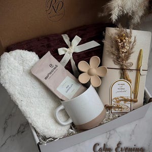May include: A gift box with a burgundy blanket, white headband, a mug with a pink base, a chocolate bar, a flower-shaped object, a notebook, and a gold pen. The box is labeled "RD Boutique" and includes the text "Calm Evening".