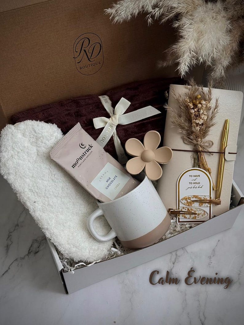 May include: A gift box with a burgundy blanket, white headband, a mug with a pink base, a chocolate bar, a flower-shaped object, a notebook, and a gold pen. The box is labeled "RD Boutique" and includes the text "Calm Evening".