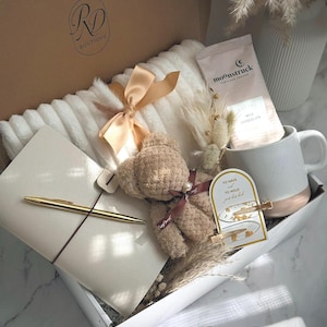 May include: A gift box with a cozy theme, containing a white blanket with a tan bow, a teddy bear, a notebook with a gold pen, a mug, and a chocolate packet. The box is labeled "RD Boutique" and has the words "Cozy Love" on the bottom.