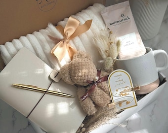 May include: A gift box with a cosy theme, containing a white blanket with a tan bow, a teddy bear, a notebook with a gold pen, a mug, and a chocolate packet. The box is labelled "RD Boutique" and has the words "Cozy Love" on the bottom.