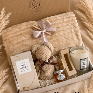 Peut inclure: Un coffret cadeau contenant une couverture beige c&ocirc;tel&eacute;e nou&eacute;e avec un ruban et un b&acirc;ton &agrave; miel en bois, un ours en peluche, une bo&icirc;te de Hot Cocoa Velvety White, une pince &agrave; cheveux beige, un petit pot de miel et une bougie Only For You.