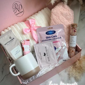 May include: A pink gift box filled with self-care items. Includes a white and beige mug, a pink bow, a white blanket, a pink towel, skincare products, and a glass jar of bath salts. The box has the text "RD Boutique" and "Luxe Moments".