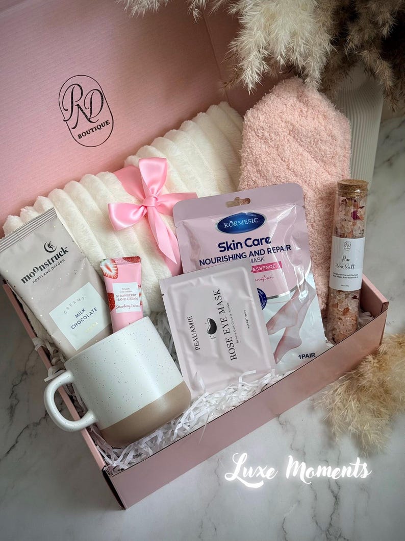 May include: A pink gift box filled with self-care items. Includes a white and beige mug, a pink bow, a white blanket, a pink towel, skincare products, and a glass jar of bath salts. The box has the text "RD Boutique" and "Luxe Moments".