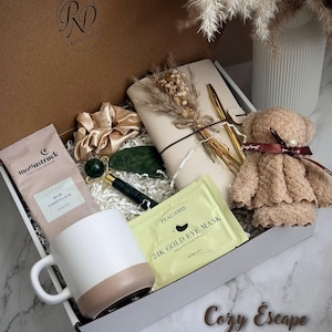 May include: A gift box filled with self-care items. Includes a white and tan mug, chocolate, a jade roller, eye mask, a teddy bear, a scrunchie, a notebook, and a pen. The box is labeled "Cozy Escape". The box has the logo "RD Boutique".