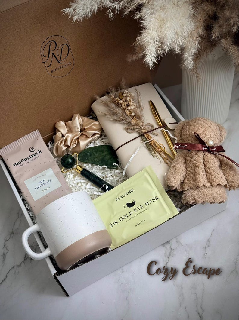 May include: A gift box filled with self-care items. Includes a white and tan mug, chocolate, a jade roller, eye mask, a teddy bear, a scrunchie, a notebook, and a pen. The box is labeled "Cozy Escape". The box has the logo "RD Boutique".