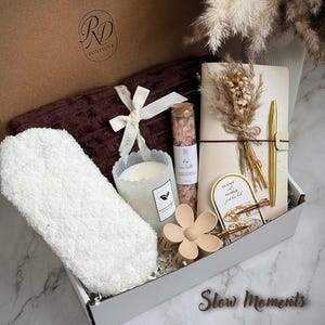 May include: A gift box with a brown lid and white interior, containing a plush white sock, a candle, a notebook, a pen, a hair clip, and a small bottle of pink salt. The box also includes a brown towel and is decorated with dried flowers. The text "Slow Moments" is visible.
