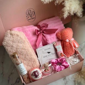May include: A pink gift box filled with self-care items. Includes a pink plush towel, a teddy bear, a bath bomb, a tube of strawberry hand cream, a scrunchie, a candle, and a Rose Eye Mask. The box is labeled "RD Boutique" and the text "Soft Blush".
