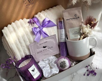 May include: A gift box filled with self-care items. Includes a white plush blanket tied with a purple ribbon, a purple candle, a mug, bath products, and a face mask. The box is labeled "Violet Dream".