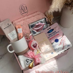 May include: A pink gift box filled with self-care items. Includes a white and brown mug, a pink towel, a lip mask, a foot mask, a bath bomb, a bar of milk chocolate, and a pair of socks. The box is tied with a pink ribbon and has the text "Moments of You".