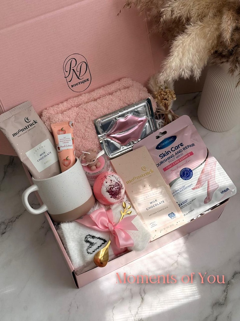 May include: A pink gift box filled with self-care items. Includes a white and brown mug, a pink towel, a lip mask, a foot mask, a bath bomb, a bar of milk chocolate, and a pair of socks. The box is tied with a pink ribbon and has the text "Moments of You".